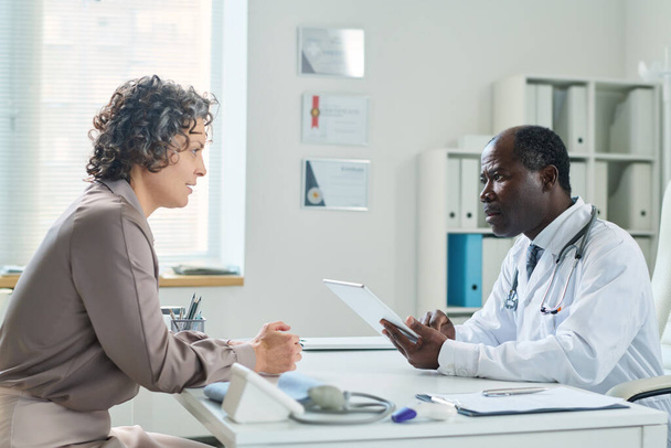 Seitenansicht eines erfahrenen Arztes mit Tablet beim Ausfüllen eines elektronischen Arztformulars, während er Patientin zuhört, die vor ihm sitzt - Foto, Bild