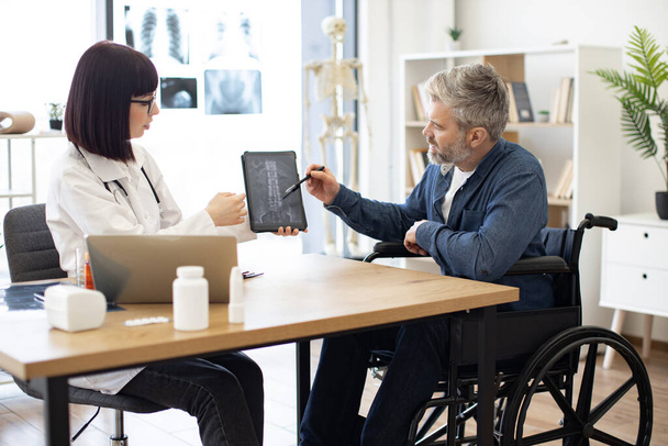 Attractive caucasian man taking pen for marking on tablet with x-ray image carried by radiologist in consulting room. Confident specialist discussing results of diagnostic tests with wheelchair user. - Foto, imagen