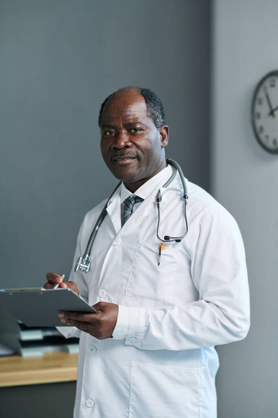 Zelfverzekerde volwassen Afro-Amerikaanse mannelijke clinicus in laboratoriumjas met stethoscoop op de hals met medisch document in functie - Foto, afbeelding