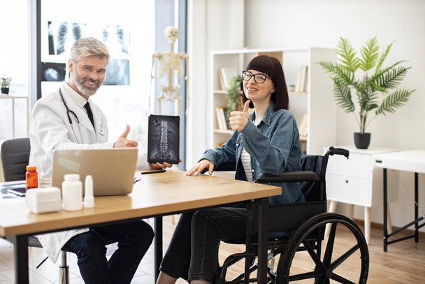 Full length portrait of friendly doctor and happy woman with disability on meeting in consulting room of clinic. Cheerful physician showing x-ray on tablet with positive results to lady in wheelchair. - Foto, afbeelding