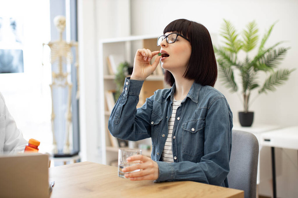 Attractive woman in everyday wear taking round pill with glass of water while sitting at office desk in modern office. Charming brunette in spectacles being prescribed antibiotics by family doctor. - Fotografie, Obrázek