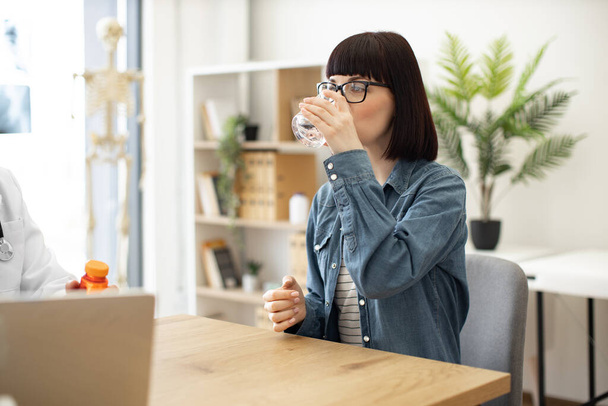 Pretty caucasian female in casual wear drinking water while seating at writing desk in doctors office of health clinic. Beautiful woman taking oral medication that being swallowed with liquid. - Foto, imagen
