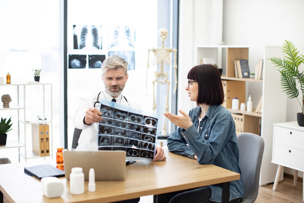 Mindful gray-haired male in lab coat holding MRI scans while brunette lady sitting next to him at desk in hospital. Family doctor examining diagnostic test results while developing treatment plan. - 写真・画像