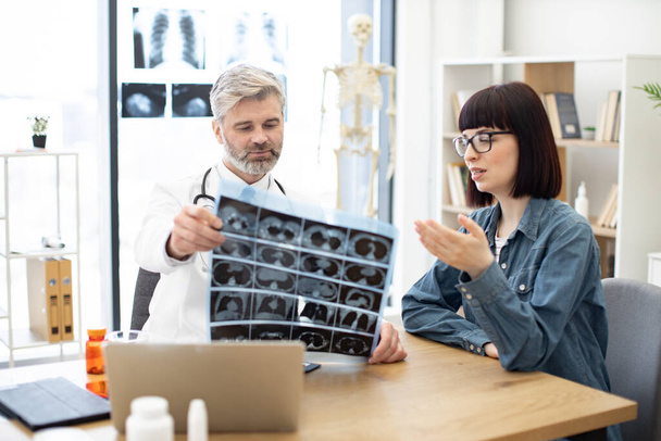 Mindful gray-haired male in lab coat holding MRI scans while brunette lady sitting next to him at desk in hospital. Family doctor examining diagnostic test results while developing treatment plan. - Foto, immagini