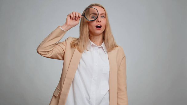 Onderzoeker onderzoeker wetenschapper kantoor vrouw houden vergrootglas in de buurt van gezicht, kijken in de camera met grote zoomde grappige ogen, zoeken analyseren. Zakelijk meisje geïsoleerd op grijze studio achtergrond - Foto, afbeelding