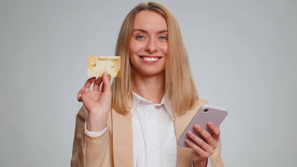 Sincere business woman customer using credit bank card and smartphone while transferring money, purchases online shopping, payment. Finance and internet. Young girl isolated alone on gray background - Foto, Bild
