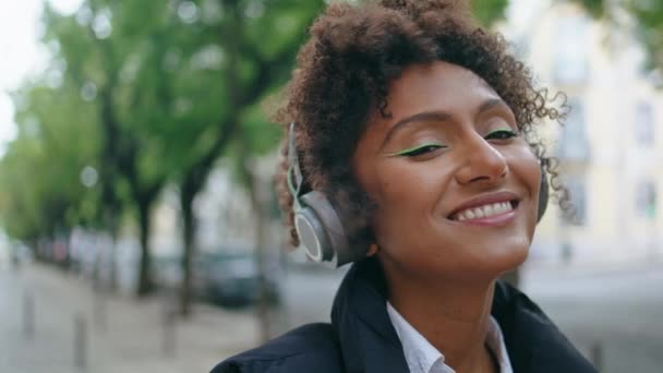 Zorgeloos Afrikaans Amerikaans meisje dat muziek luistert met een draadloze koptelefoon op straat. Vrolijk lachende vrouw die naar de camera kijkt en een moderne headset buiten draagt. mooie dame genieten van mooi geluid. - Video
