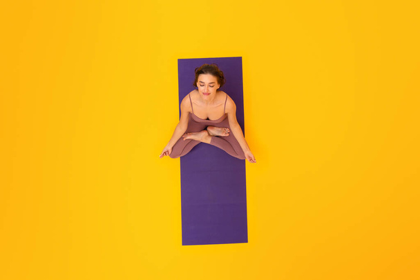 Γυμναστική Ζεν. Χαλαρωμένη γυναίκα που κάνει γιόγκα κάθεται στο Lotus Pose στο Στρώμα Γυμναστικής, χαλαρώνοντας με τα μάτια κλειστά πάνω από κίτρινο φόντο στο στούντιο. Η Λαίδη Αγκαλιάζει το μυαλό μέσω του Διαλογισμού. Πάνω όψη - Φωτογραφία, εικόνα