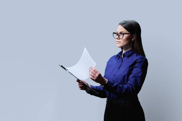 Young serious confident business woman holding clipboard with business papers documents, on grey studio background, looking in profile, copy space - Foto, afbeelding