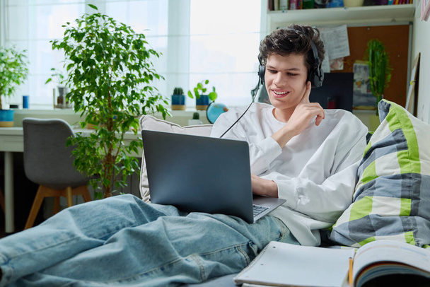 Young guy in headphones lying on the couch at home using a laptop. Smiling male studying, resting, communicating, Internet online technology, modern lifestyle - Foto, Imagen