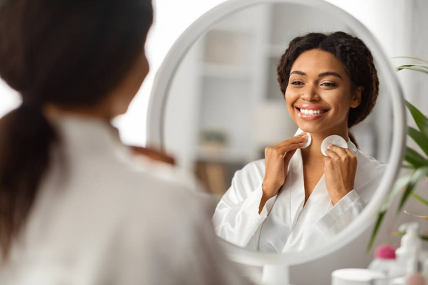 Sorrindo preto mulher limpeza pescoço pele com almofadas de algodão e olhando no espelho, bela jovem afro-americana fêmea fazendo rotina de beleza diária em casa, foco seletivo na reflexão - Foto, Imagem
