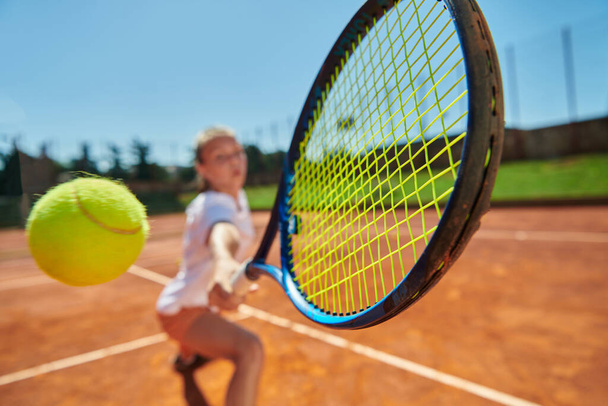 Close up photo of a young girl showing professional tennis skills in a competitive match on a sunny day, surrounded by the modern aesthetics of a tennis court - Photo, image