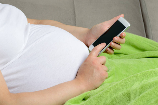 Young pregnant woman using a digital phone while relaxing on the sofa at home - Valokuva, kuva