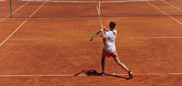 A young girl showing professional tennis skills in a competitive match on a sunny day, surrounded by the modern aesthetics of a tennis court - Photo, image