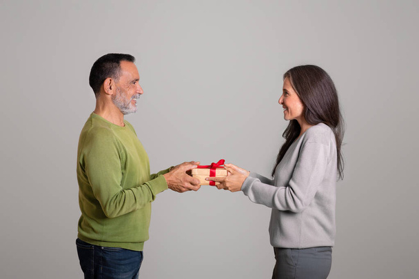 Happy caucasian aged husband gives box gift celebrating birthday, isolated on gray background, studio, profile. People emotions, congratulations on holiday, anniversary, valentine day - Foto, imagen