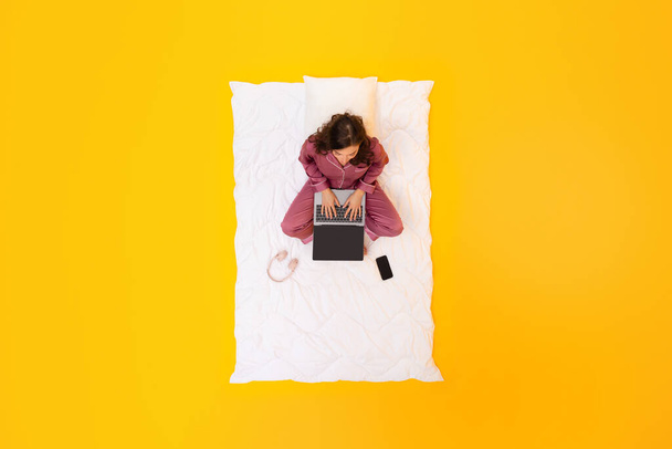 Фрилансер. Над просмотром женщины в ночном платье Многозадачность на ноутбуке работает в Интернете, прежде чем спать, сидя на одеяле над желтым фоном студии. Концепция уравновешивания трудовой жизни - Фото, изображение