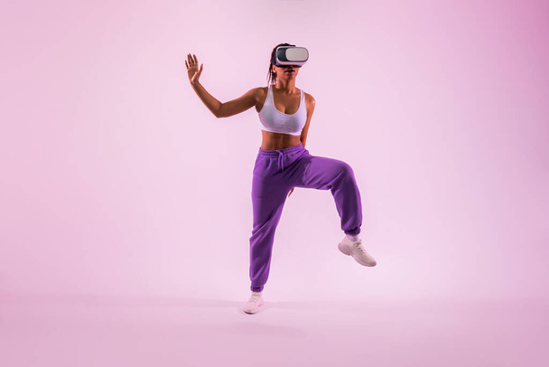 Melhorar a aptidão através da realidade vital. Mulher negra ativa com óculos VR dançando e explorando a tecnologia 3D sobre fundo de luz de néon rosa, comprimento total - Foto, Imagem