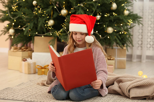 Cute little girl in Santa hat reading book near tree and gifts at home. Christmas atmosphere - Photo, image
