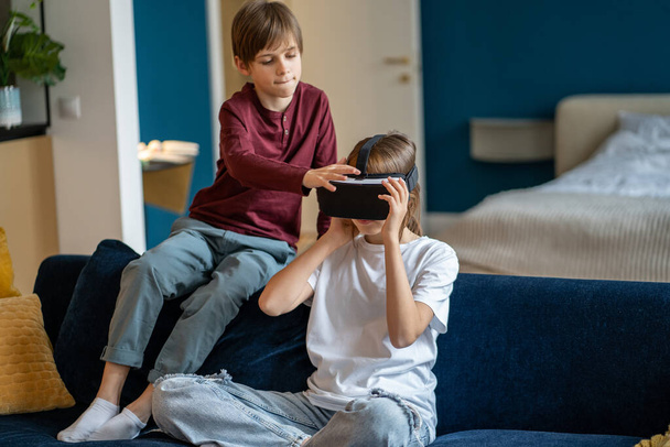 Çocuklar VR cihazını kullanırken evde birlikte vakit geçiriyorlar, çocuklar kanepede oturuyor, sanal gerçeklik kulaklığı takıyorlar, oyunlar oynuyorlar ve dünyayı keşfediyorlar. - Fotoğraf, Görsel