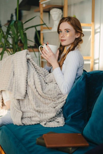 Фотографічний портрет червонуватої позитивної самотньої жінки, яка вільно сидить на дивані, покритому ковдрою під час пиття ранкового гарячого чаю. Сниться, насолоджується
 - Фото, зображення