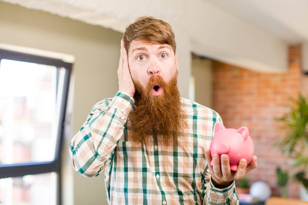 red hair man feeling extremely shocked and surprised with a piggy bank - Foto, imagen