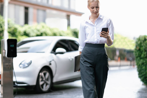 Businesswoman using tablet, walking while recharging her electric vehicle with charging station at public car parking. Progressive lifestyle of technology and ecological concern by EV car - Foto, Imagen