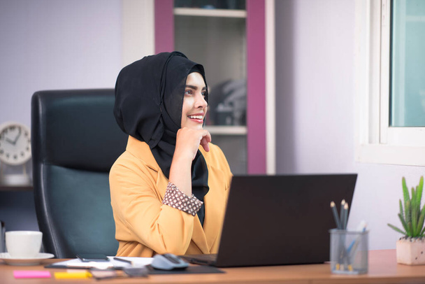 An office worker for an Asian Muslim woman sitting in front of a laptop computer at her desk and talking on a mobile phone and working at the office. - Foto, afbeelding