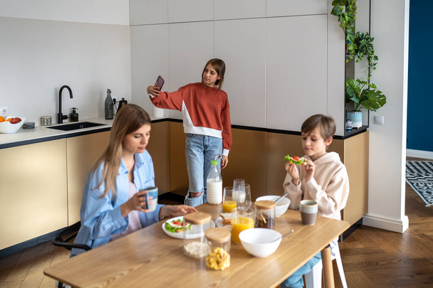 Family morning, teen daughter making selfie for social media on smartphone, mom son having breakfast sitting at table on kitchen. Motherhood, rising children, parenting, life with teenagers concept. - 写真・画像