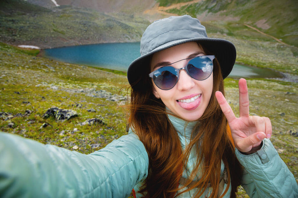Selfie portrait of young attractive woman wearing sunglasses and backpack smiling with lake and mountains in the background, sunny morning. - Foto, immagini
