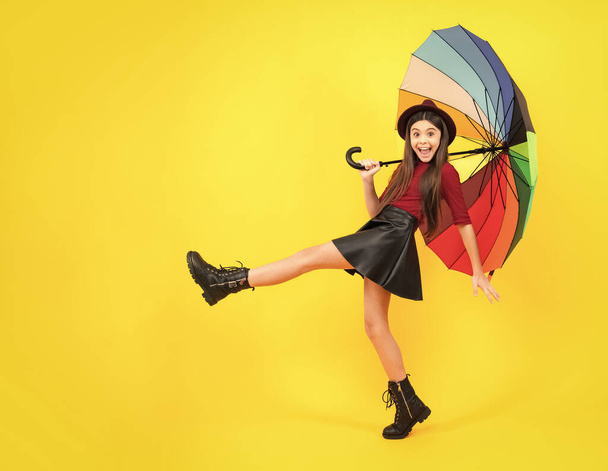 Fashion autumn kids weather. Child teenager girl with umbrella in autumn black leather skirt boots and hat isolated on yellow studio background. Excited teenager, glad amazed and overjoyed emotions - Fotoğraf, Görsel