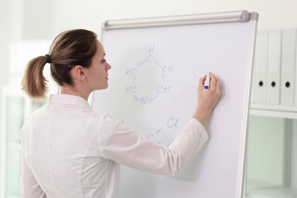 Una joven científica vestida de blanco escribe fórmulas químicas en una gran pizarra blanca intensamente. Mujer conduce seminario de química en la sala de oficina de luz - Foto, Imagen