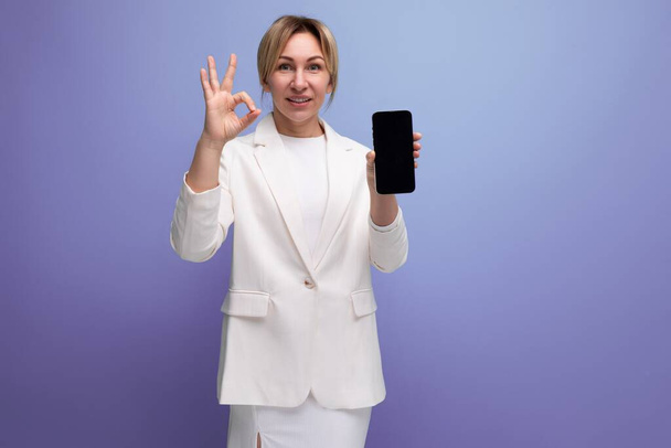 giovane donna manager bionda in una giacca elegante bianca e gonna mostra lo schermo di uno smartphone con un mockup. - Foto, immagini
