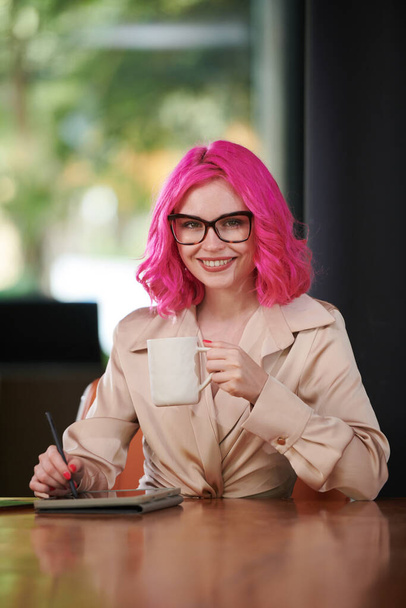 Portrait of happy young businesswoman with pink hair drinking coffee and signing documents on tablet - Foto, immagini