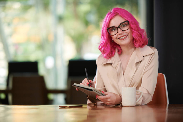 Portrait of happy young entrepreneur in glasses filling planner on tablet computer when drinking morning coffee - Photo, Image