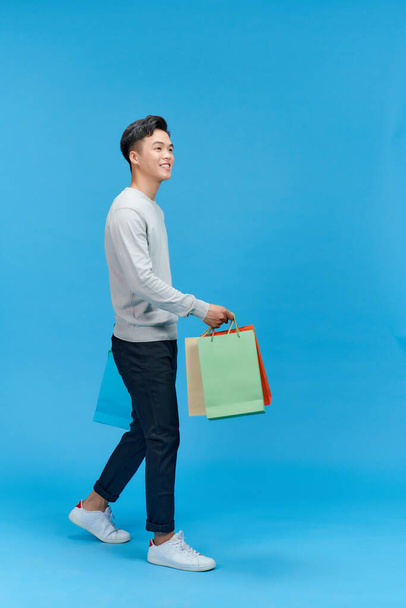 Νεαρός Ασιάτης πολύ χαρούμενος κρατώντας τσάντες για ψώνια, απομονωμένος σε μπλε φόντο. - Φωτογραφία, εικόνα