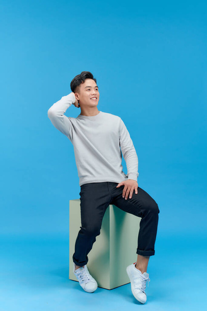 счастливый красивый молодой человек улыбается на камеру, сидя в одиночестве на голубом фоне - Фото, изображение