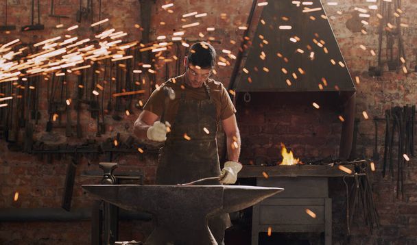 Hammer, anvil and fire with a man in a forge workshop for metal work manufacturing or production. Industry, welding and trade with a male blacksmith at work in a factory, plant or industrial foundry. - Photo, Image
