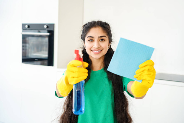 Cheerful young housewife in yellow gloves holding household chemicals and sponge in modern kitchen. - Photo, Image