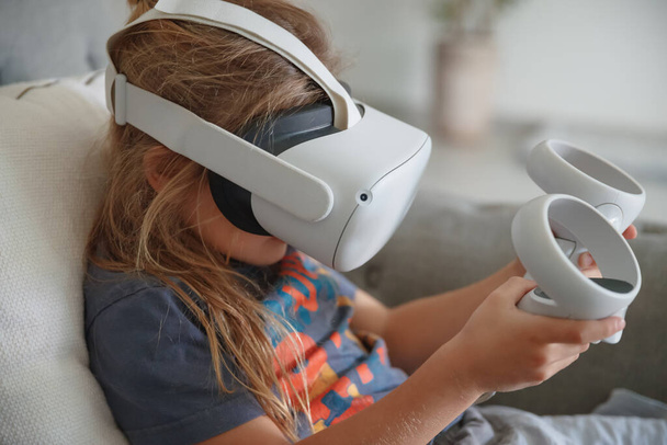 Photo of a little boy wearing VR mask playing video game at home. Modern technology. Simulation of a virtual reality. Smart child enjoying cyberspace. - Photo, image