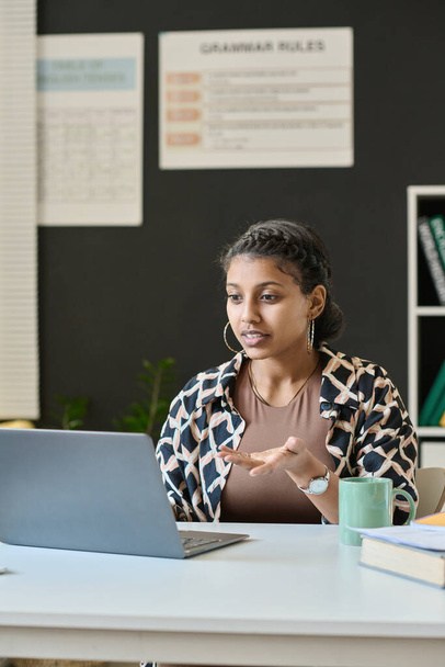 Vertical image of student sitting at desk in front of laptop and talking to teacher online on laptop - Fotoğraf, Görsel