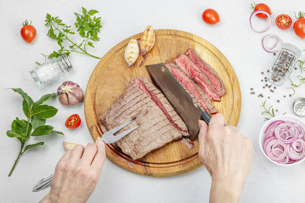 Woman cuts grilled steak. Female hands with knife and two pronged fork. Fresh herbs, spices, vegetables. Wooden cooking board, picnic or barbecue concept. Light stone concrete background, top view - Photo, Image