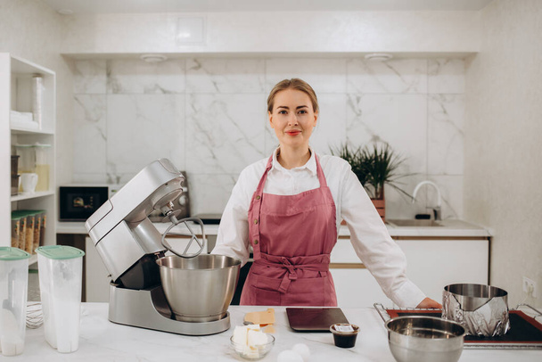 Hermosa pastelera femenina prepara dulce en la cocina moderna mirando a la cámara - Foto, imagen
