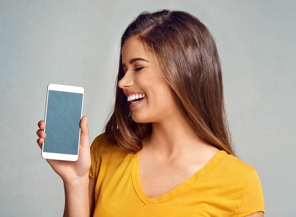 Youll definitely want to check this out. Studio shot of an attractive young woman holding a cellphone with a blank screen against a grey background - Photo, Image