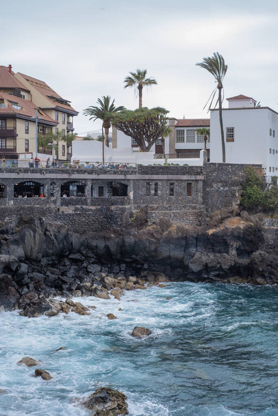 Näkymä taloihin vulkaanisella kalliolla. Aallot murtautuvat jyrkänteen vulkaanisia kiviä vasten. Rankka meri. Mustia kiviä. Puerto de la Cruz, Tenerife, Kanariansaaret, Espanja. - Valokuva, kuva