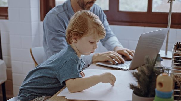 Отец работает из домашнего офиса с помощью ноутбука сидеть за столом с милым маленьким сыном играет почти нарушить. - Фото, изображение