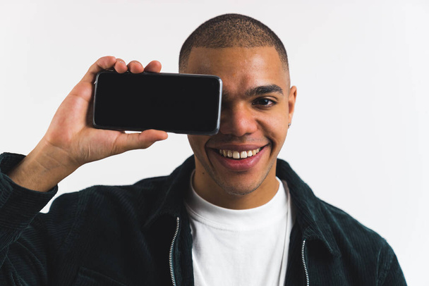 Χαρούμενος Αφρο-Αμερικάνος που κρατάει ένα τηλέφωνο οριζόντια κοντά στο πρόσωπό του, άδεια οθόνη, μεσαίο κοντινό πλάνο. Υψηλής ποιότητας φωτογραφία - Φωτογραφία, εικόνα