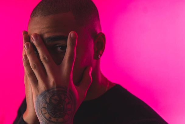 closeup shot of a young tattooed man covering his face with hands and looking to the camera, pink background. High quality photo - Fotoğraf, Görsel
