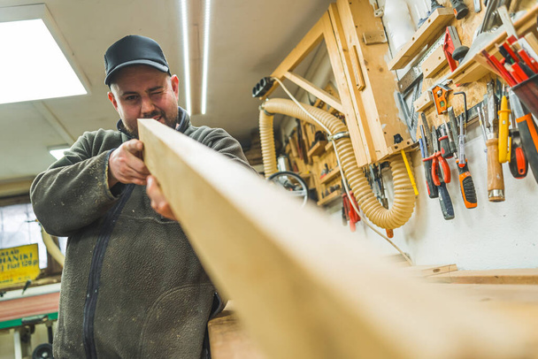 Ξυλουργός εξετάζει μια ξύλινη σανίδα μετά την ισοπέδωση χρησιμοποιώντας μηχανή ξύλου. Υψηλής ποιότητας φωτογραφία - Φωτογραφία, εικόνα