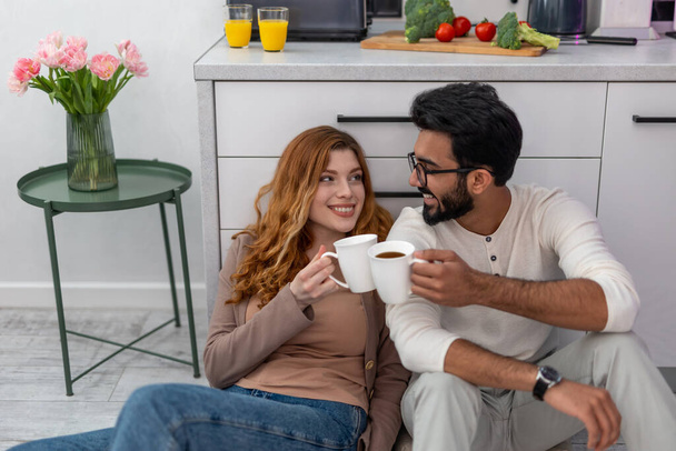 Пара, сидящая на полу на кухне и разговаривающая, пьющая утренний кофе дома. - Фото, изображение