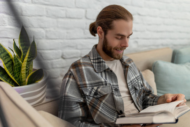 Handsome smiling happy young man reading book while sitting on sofa in his living room. - Photo, Image